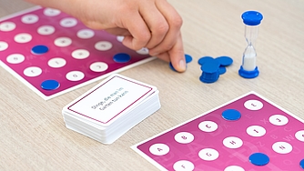 Hand legt blauen Spielstein auf Spielplan mit Buchstabenfeldern und Sanduhr daneben